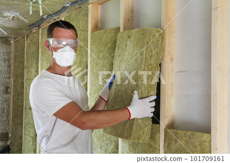 Worker in protective goggles and respirator insulating rock wool insulation in wooden frame for future house walls for cold barrier. Comfortable warm home, economy, construction and renovation concept 101709161