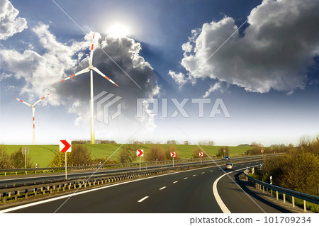 Wide highway with moving car stretching to horizon by green hills with high wind turbines on blue cloudy sky background. Modern technologies, renewable energy source and ecological problems solving. Wide highway with moving car stretching to horizon by green hills with high wind turbines on blue cloudy sky background. Modern technologies, renewable energy source and ecological problems solving. 101709234