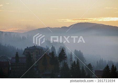 Resort village houses buildings on background of foggy blue mountain hills covered with dense misty spruce forest under bright pink sky at sunrise. Mountain landscape at dawn. Resort village houses buildings on background of foggy blue mountain hills covered with dense misty spruce forest under bright pink sky at sunrise. Mountain landscape at dawn. 101711034
