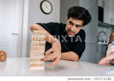 Portrait of skilled concentrated young man carefully removing wooden cube from tower structure while enjoying fun leisure time activity. Cheerful people having fun and enjoying competition. Portrait of skilled concentrated young man carefully removing wooden cube from tower structure while enjoying fun leisure time activity. Cheerful people having fun and enjoying competition. 101711280