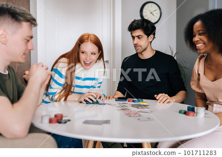 Happy overjoyed young woman laughing celebrating victory in game of poker sitting at table in living room. Group of multiethnic men and women having fun and enjoying competition at home. 101711283