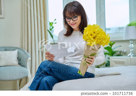 Happy middle aged female reading congratulations note, with bouquet of flowers, at home Happy middle aged female reading congratulations note, with bouquet of flowers, at home 101711736