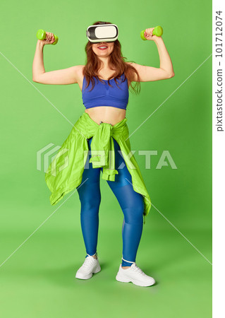 One young charming girl, woman wearing vr glasses doing workout with dumbbells and smiling at camera over green background. Girls power 101712074