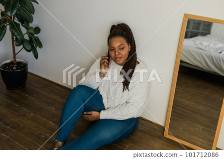 Positive young african american woman chatting on cellphone and smiling on floor at home. Cheerful young woman communicating with someone on smartphone indoors Positive young african american woman chatting on cellphone and smiling on floor at home. Cheerful young woman communicating with someone on smartphone indoors 101712086