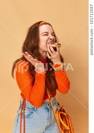 Portrait of one beautiful young girl wearing orange clothes eating donut with pleasure and closed eyes over ginger studio background. Sweettooth Portrait of one beautiful young girl wearing orange clothes eating donut with pleasure and closed eyes over ginger studio background. Sweettooth 101712087