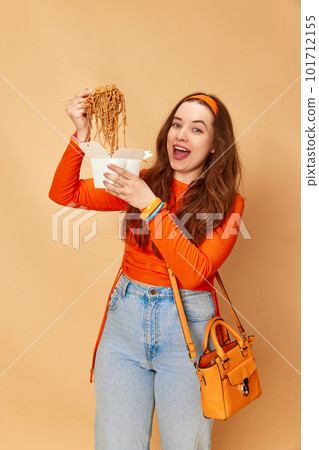 Portrait with young charming girl holding box with food and eating noodles with hunger face on ginger studio background. Junk, unhealthy food Portrait with young charming girl holding box with food and eating noodles with hunger face on ginger studio background. Junk, unhealthy food 101712155
