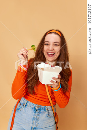 Portrait of pretty girl, student holding box with food and eating broccoli with smile on ginger studio background. Healthy menu for a day Portrait of pretty girl, student holding box with food and eating broccoli with smile on ginger studio background. Healthy menu for a day 101712197