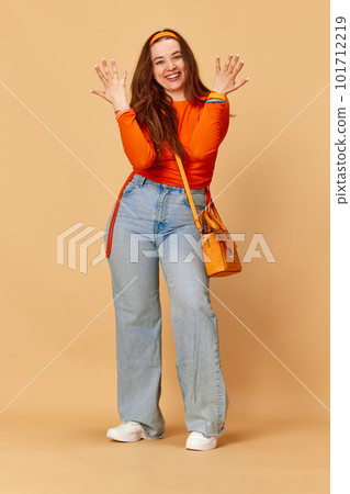 Portrait of adorable girl, student wearing bright clothes and jeans showing palms and smiling over ginger studio background. Young hippie girl Portrait of adorable girl, student wearing bright clothes and jeans showing palms and smiling over ginger studio background. Young hippie girl 101712219