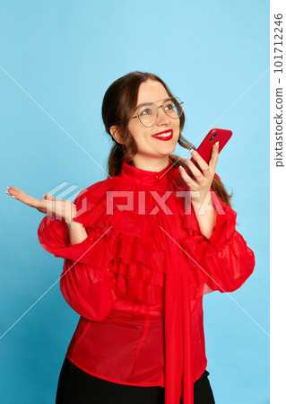 One young beautiful girl, student wearing eyeglasses talking via cell phone over blue studio background. Online communication One young beautiful girl, student wearing eyeglasses talking via cell phone over blue studio background. Online communication 101712246