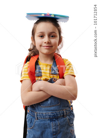 Authentic portrait Caucasian 5-6 years old positive cute child girl, first grader with book on her head, smiling cutely, looking at camera, posing with her arms crossed over isolated white background 101712854