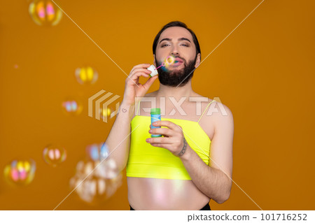 Playful bearded transsexual person wearing yellow top blowing soap bubbles having fun 101716252