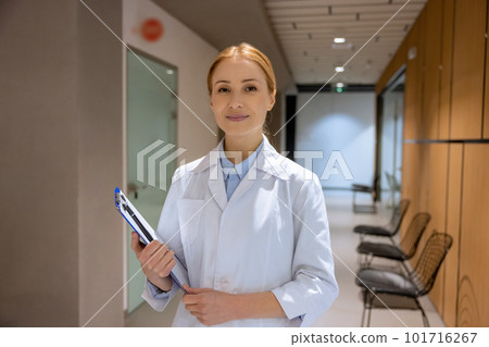 Attractive blonde young female doctor standing in hospital corridor and holding clipboard. 101716267