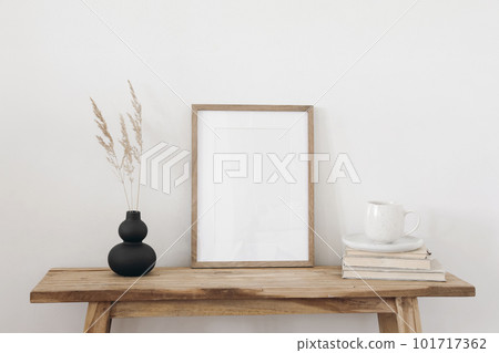 Blank vertical wooden picture frame mockup. Organic shaped black vase with dry grass on table, bench. Cup of coffee on old books. Working space, home office. Art, poster display. Boho beige interior. Blank vertical wooden picture frame mockup. Organic shaped black vase with dry grass on table, bench. Cup of coffee on old books. Working space, home office. Art, poster display. Boho beige interior. 101717362
