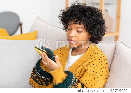 African american woman shopping online holding smartphone paying with gold credit card. Girl sitting at home buying on Internet enter credit card details. Online shopping ecommerce delivery service African american woman shopping online holding smartphone paying with gold credit card. Girl sitting at home buying on Internet enter credit card details. Online shopping ecommerce delivery service 101719011