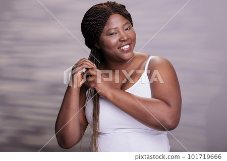 Body positive woman styling hair and doing hair care routine portrait. Young african american curvy model looking at camera and making braids in studio for haircare commercial 101719666