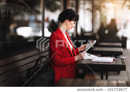 A attractive businesswoman in corporate outfit red blazer holding morning newspaper while sitting on cafe terrace. Manager is reading the latest world news during a coffee break at work A attractive businesswoman in corporate outfit red blazer holding morning newspaper while sitting on cafe terrace. Manager is reading the latest world news during a coffee break at work 101719677