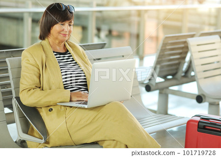 smiling adult businesswoman is working on laptop in airport terminal hall and waiting for flight. Middle ages female in yellow suit with red luggage suitcase in business trip on train or bus station smiling adult businesswoman is working on laptop in airport terminal hall and waiting for flight. Middle ages female in yellow suit with red luggage suitcase in business trip on train or bus station 101719728