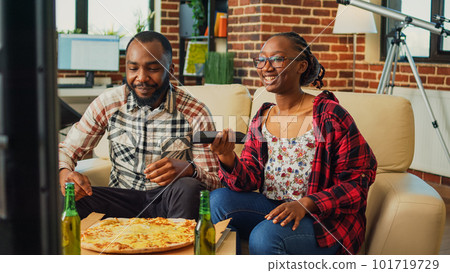 Boyfriend and girlfriend enjoying pizza slices and binge watching show in living room, eating fast food meal. Young couple receiving takeout food order and sitting on couch to have dinner. Boyfriend and girlfriend enjoying pizza slices and binge watching show in living room, eating fast food meal. Young couple receiving takeout food order and sitting on couch to have dinner. 101719729
