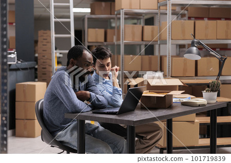Storehouse workers checking orders logistics on laptop computer before start putting trendy clothes in cardboard box, discussing packaging distribution. Diverse employees working in warehouse Storehouse workers checking orders logistics on laptop computer before start putting trendy clothes in cardboard box, discussing packaging distribution. Diverse employees working in warehouse 101719839