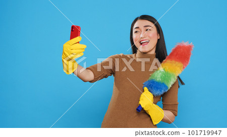 Smiling housekeeper holding colorful feather dust while taking selfie with mobile phone, standing in studio over blue background. Cheerful maid using high-quality detergents and rubber gloves 101719947