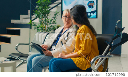 Female doctor taking notes on tablet at consultation with asian patient suffering from physical disability. Person with crutches talking to medic at checkup examination appointment. 101719990