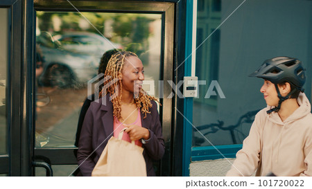 Female carrier giving fastfood package order to woman at office building front door, express delivery. Female client receiving restaurant food in paperbag from courier. Handheld shot. 101720022