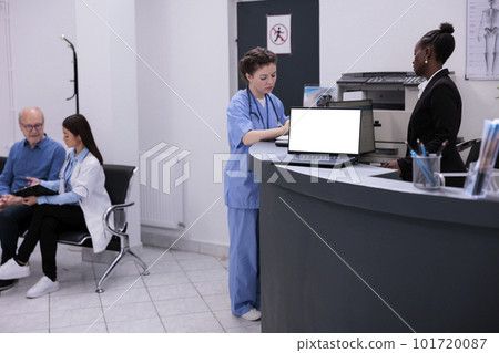 Reception worker talking with assistant discussing medical report during consultation in hospital waiting room. Laptop computer with white isolated screen standing on registration counter desk Reception worker talking with assistant discussing medical report during consultation in hospital waiting room. Laptop computer with white isolated screen standing on registration counter desk 101720087