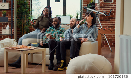 Cheerful people clinking bottles of beer and watching movie on tv channel program, having fun together at house party. Friends drinking alcohol while they watch film on television. 101720186