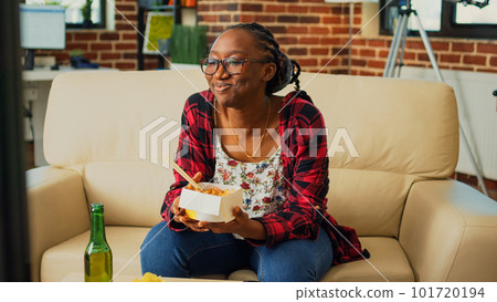 African american girl eating noodles with chopsticks, serving beer and binge watching favorite tv show on sofa. Happy woman enjoying asian meal from takeout delivery order, watch television. African american girl eating noodles with chopsticks, serving beer and binge watching favorite tv show on sofa. Happy woman enjoying asian meal from takeout delivery order, watch television. 101720194