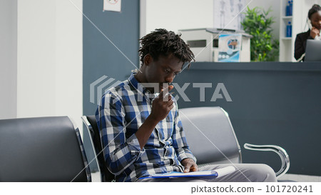 African american patient filling in checkup report papers, sitting in hospital waiting room lobby. Male person reading medical form files before attending appointment with general practitioner. 101720241