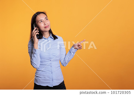 Filipino woman talking at mobile phone with remote friend, enjoying conversation in studio over yellow background. Attractive young adult answering smartphone call. Communication concept 101720305