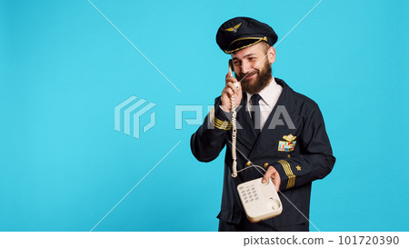 Commercial pilot talking on landline phone call in studio, answering office telephone with cord for remote conversation. Young smiling airliner in uniform chatting on phone line. 101720390