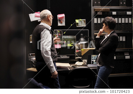 Private investigators looking at evidence board, working overhours at criminal case in arhive room. Criminology department employee discussing missing person report, planning solution to catch suspect 101720423