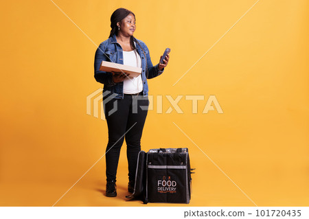African american pizzeria courier holding pizza cardboard box while checking client adreess on smartphone before delivery order during lunch time. Food transportation service and takeaway concept African american pizzeria courier holding pizza cardboard box while checking client adreess on smartphone before delivery order during lunch time. Food transportation service and takeaway concept 101720435