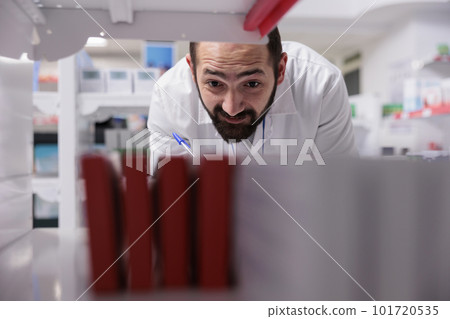 Pharmacist looking at shelves full with pills packages doing inventory writing medication name on papers, working in pharmacy. Drugstore worker is responsible for reviewing orders to ensure accuracy. 101720535