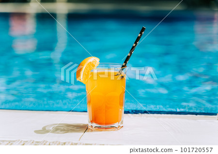 Tropical sparkling cocktail by the pool. The shot of glass with orange lemonade fruit cocktail standing near the poolside. Summer alcohol free drink by the hotel pool. Hello summer holiday vacation Tropical sparkling cocktail by the pool. The shot of glass with orange lemonade fruit cocktail standing near the poolside. Summer alcohol free drink by the hotel pool. Hello summer holiday vacation 101720557