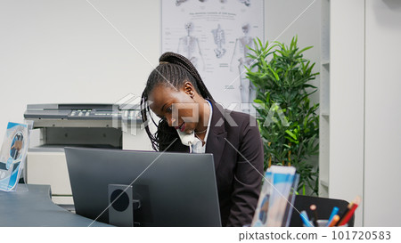 Young health center employee working at reception desk, answering landline phone call to make appointments for patients. Female receptionist using registration papers at counter, waiting room. 101720583