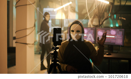 Skilled cyber criminal with white mask recording live ransomware video on dark web, making threats after stealing passwords. Masked hacker acting mysterious on camera at night, harassment. Skilled cyber criminal with white mask recording live ransomware video on dark web, making threats after stealing passwords. Masked hacker acting mysterious on camera at night, harassment. 101720618