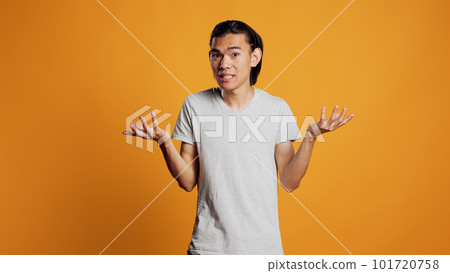 Clueless young person doing i dont know sign in studio, acting uncertain and unsure about answer to question. Asian guy being carefree and casual, acting doubtful over orange backdrop. 101720758