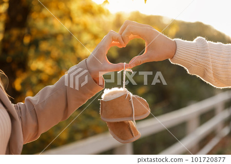 Man and pregnant wife are doing heart gesture with hands and holding warm baby shoes on nature autumn background. Future parents waiting for the baby. The concept of Mother's Day and Women's Day. Man and pregnant wife are doing heart gesture with hands and holding warm baby shoes on nature autumn background. Future parents waiting for the baby. The concept of Mother's Day and Women's Day. 101720877