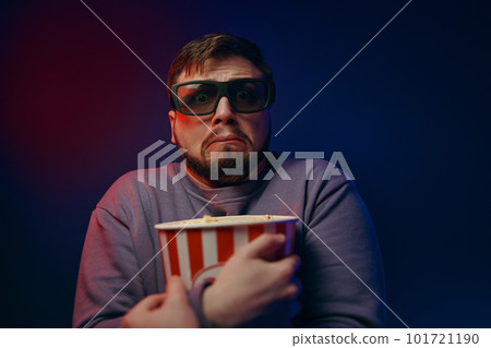 A man is in a dark room with neon lighting, watching a movie with 3D glasses and popcorn. 101721190