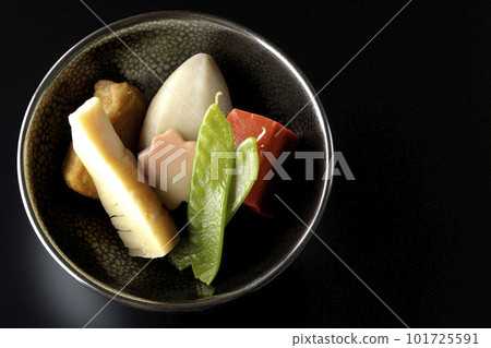 A bird's-eye view of a simmered dish containing bamboo shoots, taro, namafu, snow peas, etc. against a black background 101725591