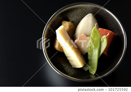 A bird's-eye view of a simmered dish containing bamboo shoots, taro, namafu, snow peas, etc. against a black background 101725592
