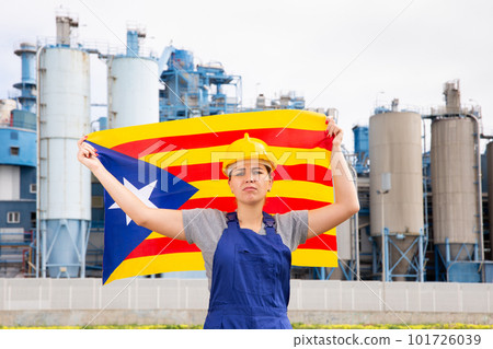 Upset disappointed young female engineer in helmet waving state flag of Estelada during strike in front of big tanks at chemical plant 101726039