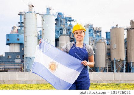 Young female engineer in helmet waving state flag of Argentina while standing in front of big tanks at chemical plant 101726051