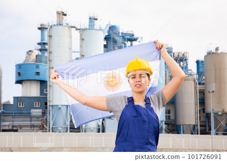 Upset disappointed young female engineer in helmet waving state flag of Argentina during strike in front of big tanks at chemical plant Upset disappointed young female engineer in helmet waving state flag of Argentina during strike in front of big tanks at chemical plant 101726091