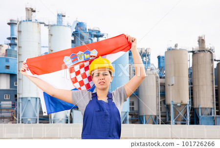 Upset disappointed young female engineer in helmet waving state flag of Croatia during strike in front of big tanks at chemical plant 101726266