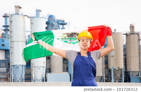 Upset disappointed young female engineer in helmet waving state flag of Mexico during strike in front of big tanks at chemical plant 101726312