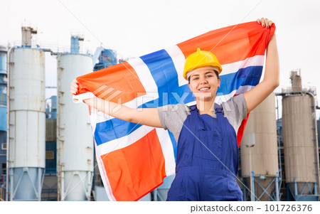 Young female engineer in helmet waving state flag of Norway while standing in front of big tanks at chemical plant 101726376