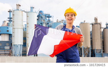 Positive young female worker in helmet waving national flag of France while standing in front of big tanks at refinery factory on sunny summer day 101726415